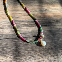 Close-up detail of Riviera Romance choker showing colorful tourmaline beads in pink, green, yellow and blue with gold accents and pearl drop pendant on rustic wooden surface