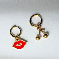 Sweet Cherries Mismatched Charm Hoop Earrings by Shop Lucky Charms — gold huggie hoops with a red enamel lip charm and a gold cherry charm, laid flat on a light grey surface