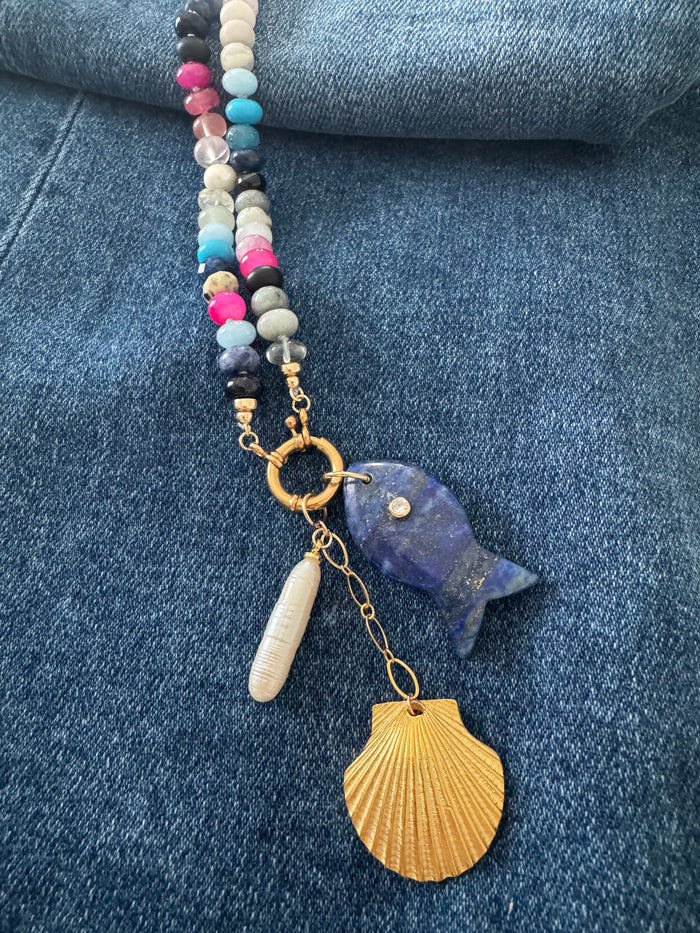 Close-up detail of Tempest Necklace showing lapis lazuli fish charm, scallop shell pendant, and mixed gemstone beads with gold toggle clasp on denim