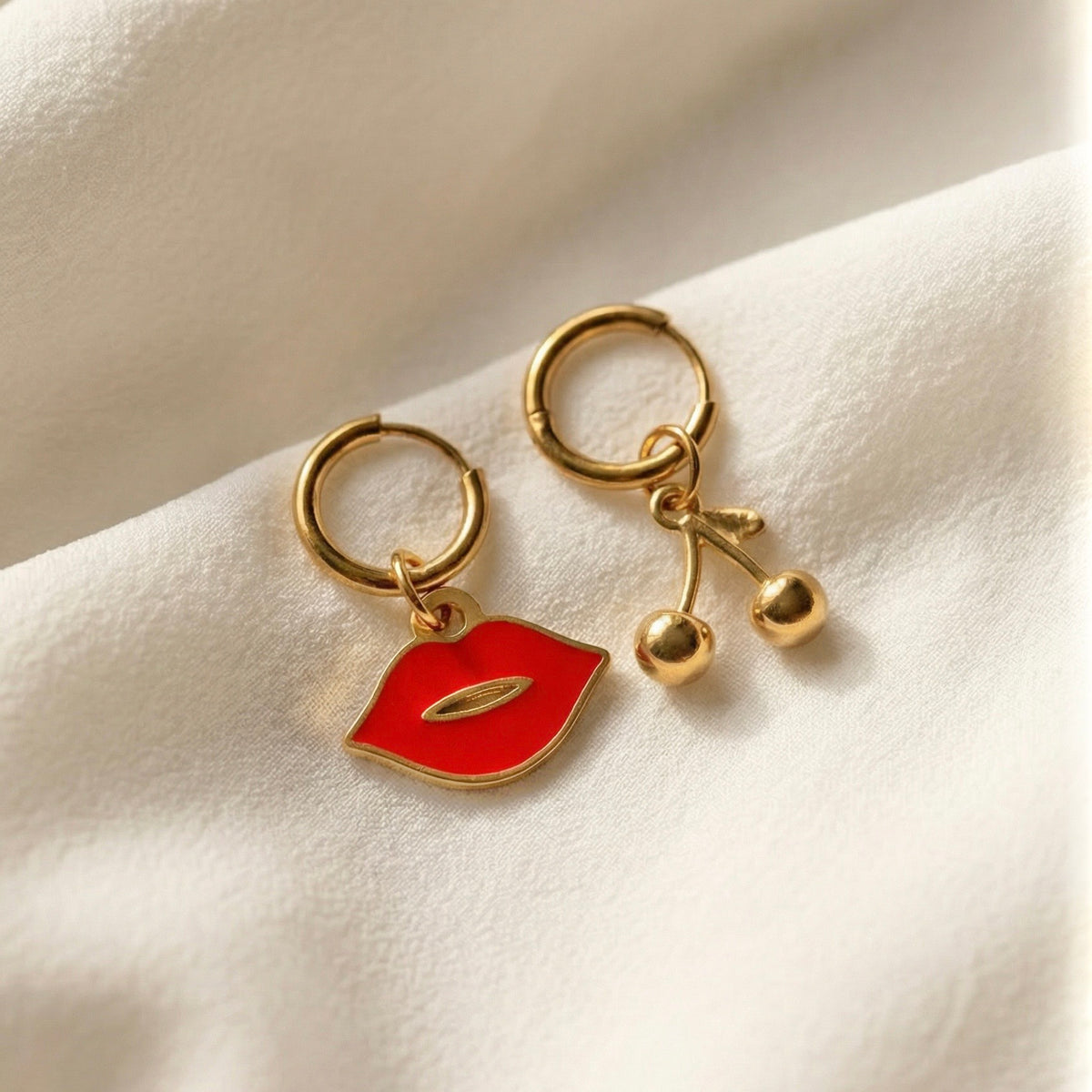 Shop Lucky Charms Sweet Cherries Mismatched Earrings — gold huggie hoops featuring a red enamel lip charm and 3D gold cherry charm styled on cream satin fabric