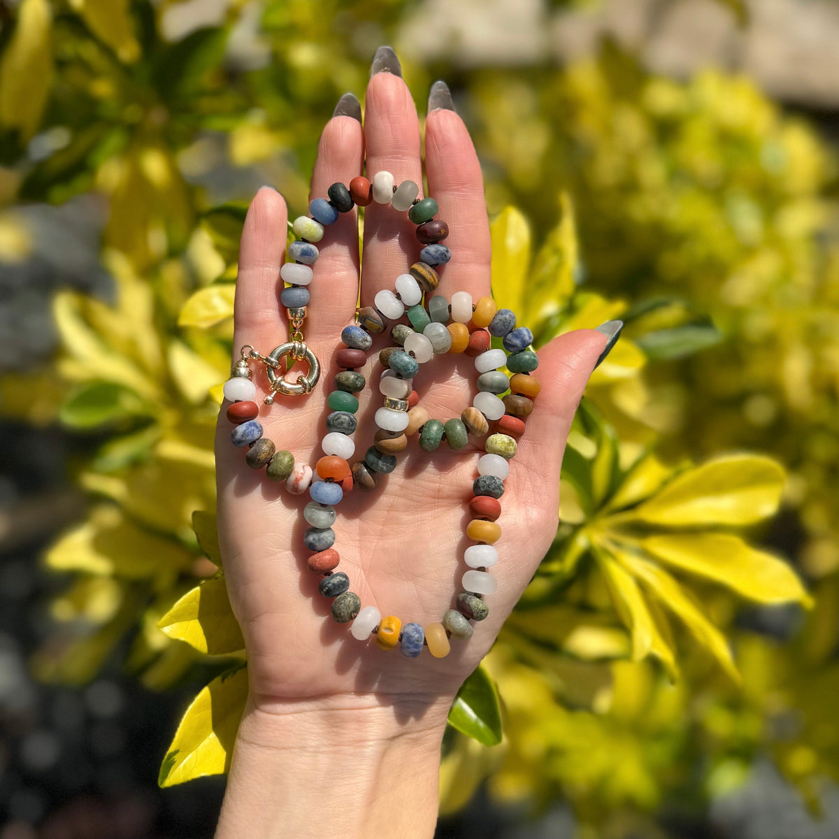 Handmade unisex gemstone necklace held in palm showing full length with matte earth tone beads and gold accents against greenery