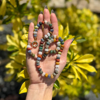 Handmade unisex gemstone necklace held in palm showing full length with matte earth tone beads and gold accents against greenery